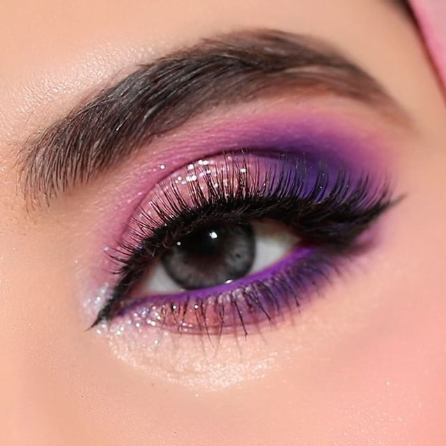 Close-up of vibrant purple eye makeup trends featuring a cut crease with soft pink blending, glittery eyelids, purple waterline eyeliner, and voluminous false lashes.