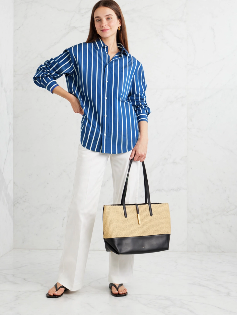 Woman wearing an oversized blue and white striped button-down shirt with white wide-leg trousers and a straw tote, showcasing chic spring outfit ideas for women.   
