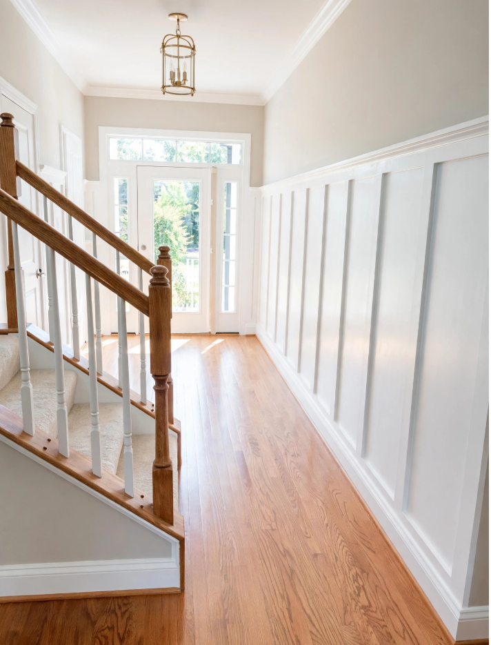 Wainscoting and Beadboard Styles: What’s the Difference? Bright entryway featuring tall white board and batten wainscoting hallway paneling, oak wood floors, and a staircase.